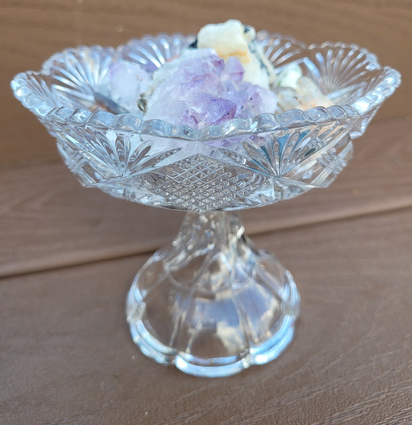 A *Vintage EAPG Pedestal Pressed Glass Dish* from *The Crystalary*, reminiscent of an ornate glass dish, holds a collection of colorful, small crystalline rocks. The clear, intricately patterned glass features a diamond and fan design. The twisted pedestal adds a unique touch as the bowl sits elegantly on a wooden surface.