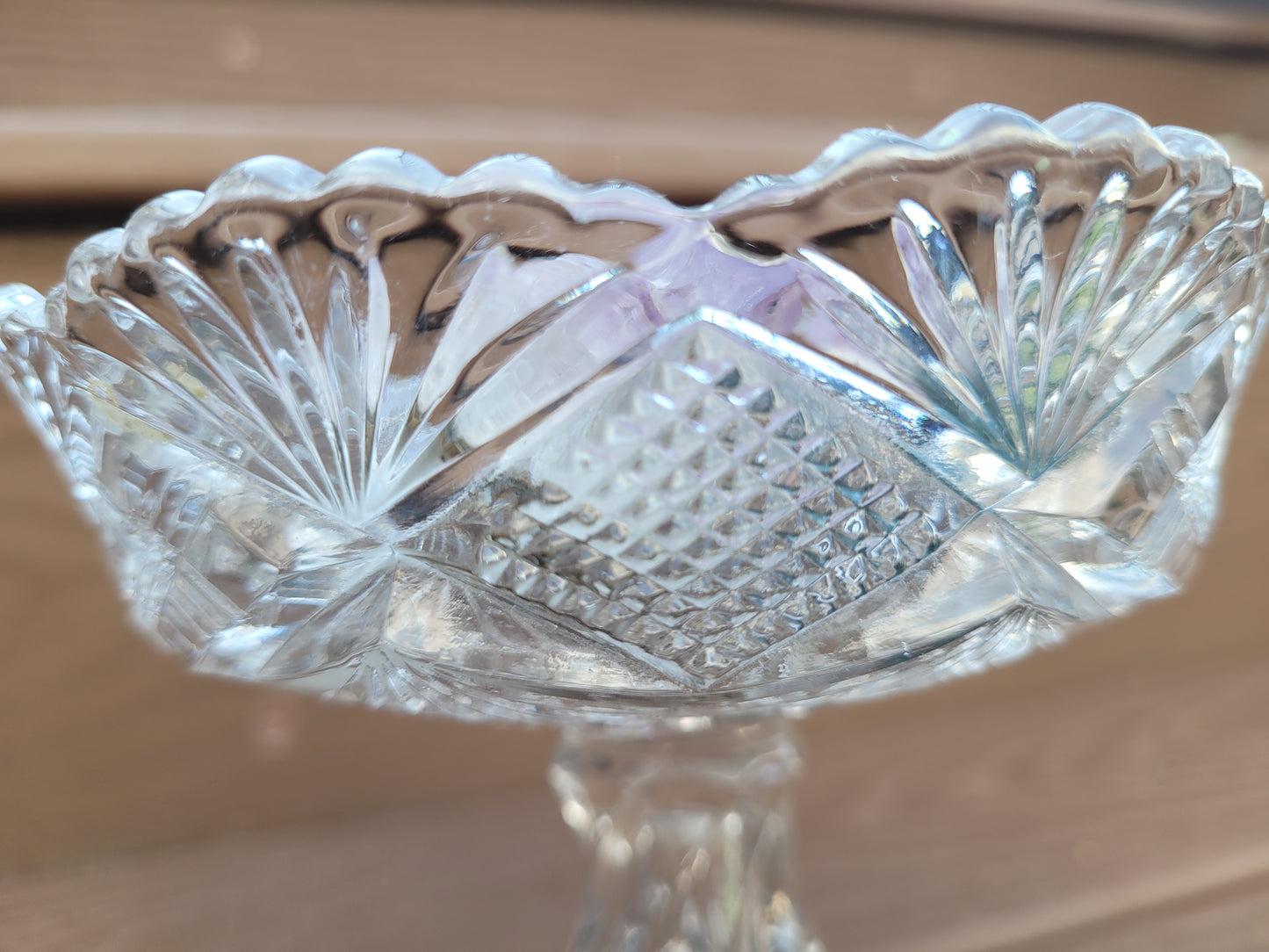 Close-up of a clear, intricately designed Vintage EAPG Pedestal Pressed Glass Dish from The Crystalary, featuring a cut glass pattern. The bowl showcases detailed starburst-like and diamond-shaped designs and is elevated on a twisted pedestal base. The background shows a blurred wooden surface, making it perfect for holding candy.