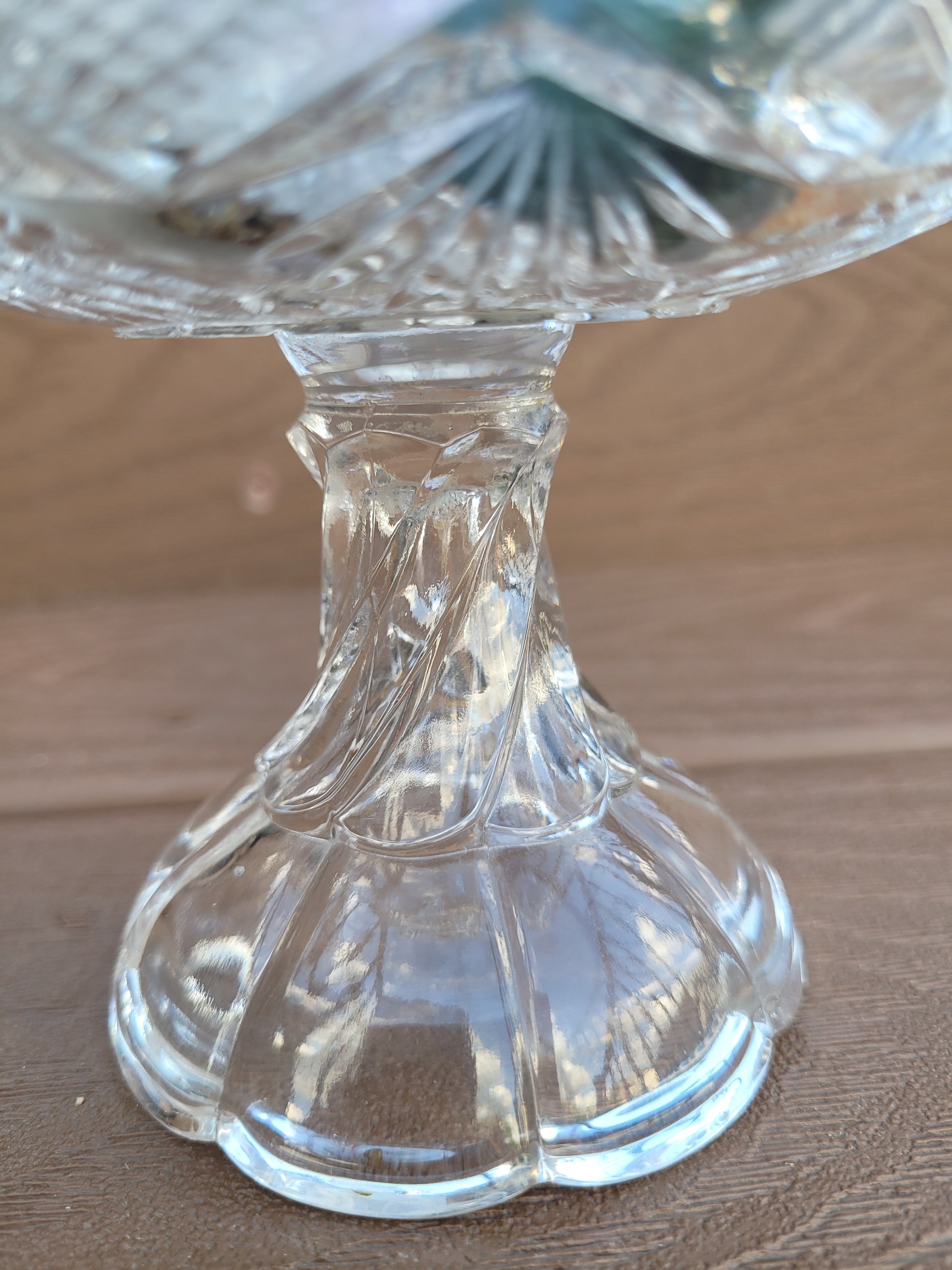 A close-up photograph of the pedestal base of The Crystalary's Vintage EAPG Pedestal Pressed Glass Dish. The twisted pedestal design rests on a wooden surface, capturing light reflections and fine details in the craftsmanship, making it perfect for showcasing candy.