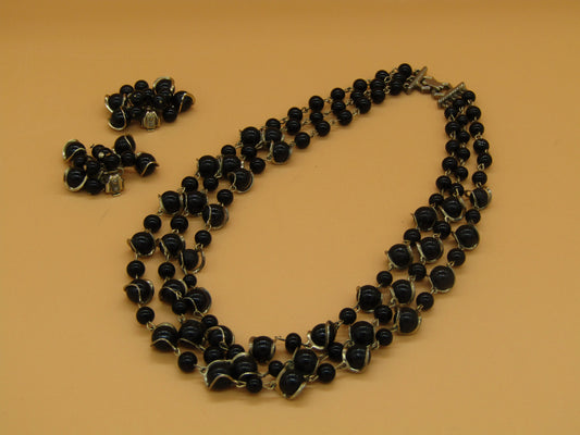 The Crystalary's Vintage Gold Tone and Black Beaded 3 Strand Necklace and Clip On Earring Set is showcased on a beige background. To the left, the matching clip-on earrings feature clusters of the same elegant black beads bordered in gold.