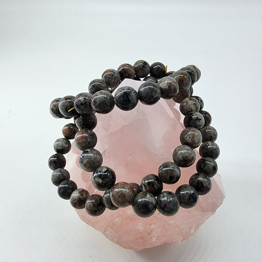 A stretch Yooperlite Crystal Bracelet by The Crystalary, made of round, dark-colored beads, is wrapped around a large, translucent pink crystal. The bracelet showcases polished spherical stones in varying shades of dark gray and brown. Under a bright orange UV reaction, this stunning piece aims to reduce inflammation.