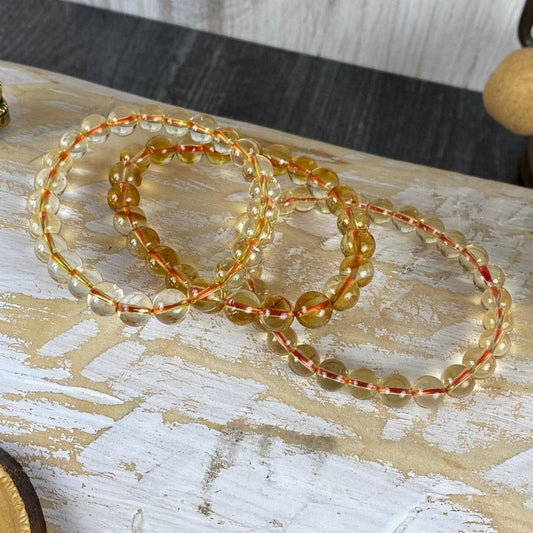 Close-up of three Citrine Crystal Bracelets by The Crystalary, arranged on a whitewashed wooden surface. The elastic bands feature both clear and amber-colored beads. Surrounding the bracelets are various decorative items including a small statue, a wooden rune, and a tasseled cloth—perfect for your new journey.