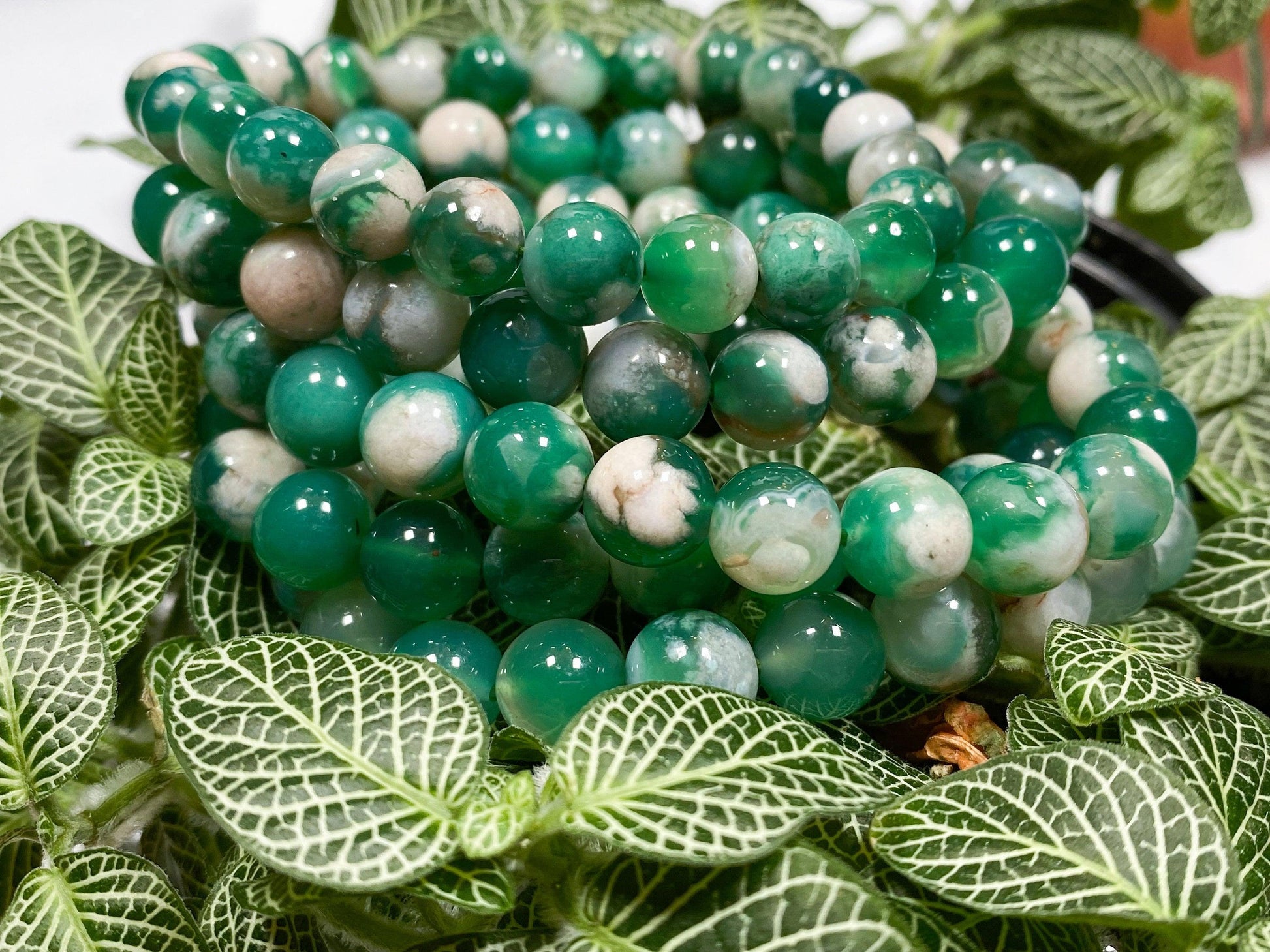 A close-up of The Crystalary’s Green Flower Agate Crystal Bracelet, featuring intricately stacked polished beads in various shades of green with white marbling, atop vibrant green and white patterned leaves. The smooth, round beads blend harmoniously with the leaves beneath, making it perfect for balancing the heart chakra.