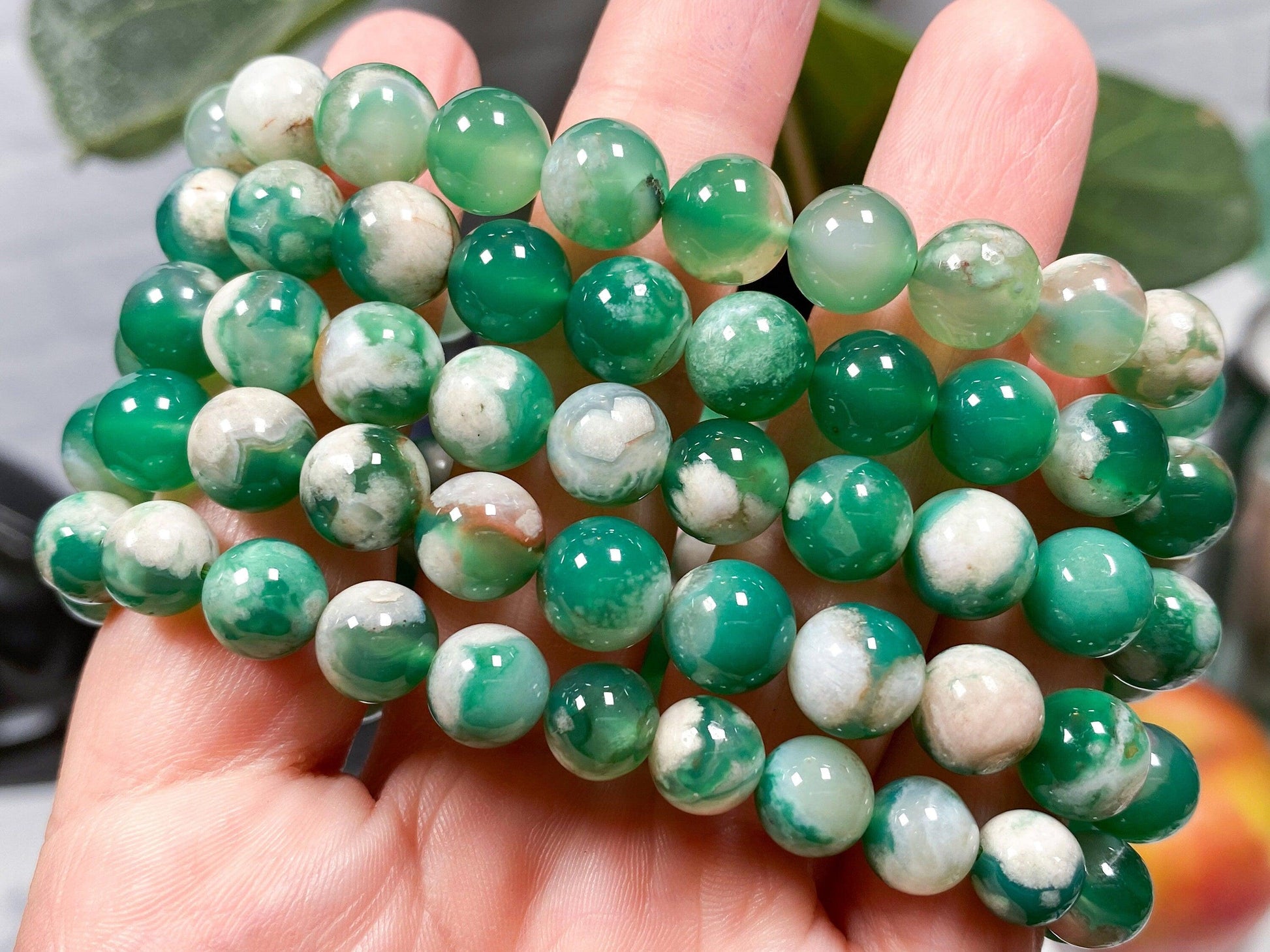 A hand holds a Green Flower Agate Crystal Bracelet by The Crystalary, showcasing several spherical beads in various shades of green, white, and translucent tones that create a marbled effect. In the out-of-focus background, a plant enhances the natural ambiance, ideal for heart chakra alignment.