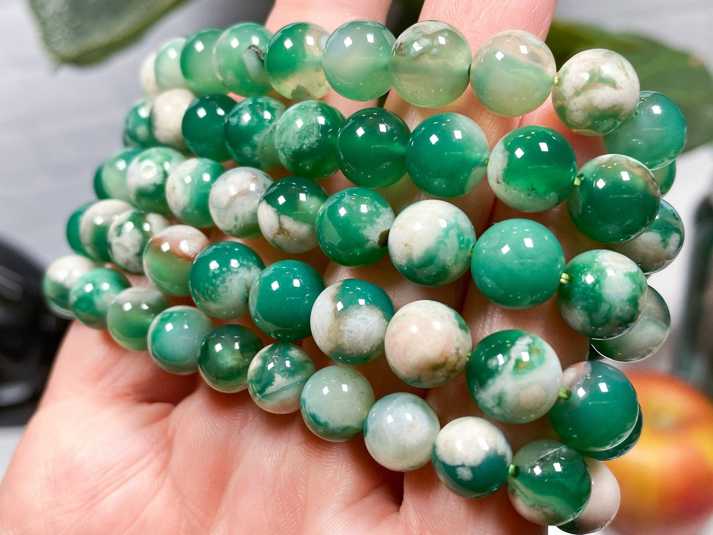 A close-up of a person's hand holding several strands of The Crystalary's Green Flower Agate Crystal Bracelet. These round, polished beads, often used for balancing the heart chakra, exhibit slight variations in shade and pattern, showcasing vibrant green hues and soft white marbling. The background is slightly blurred to emphasize the beauty of the beads.