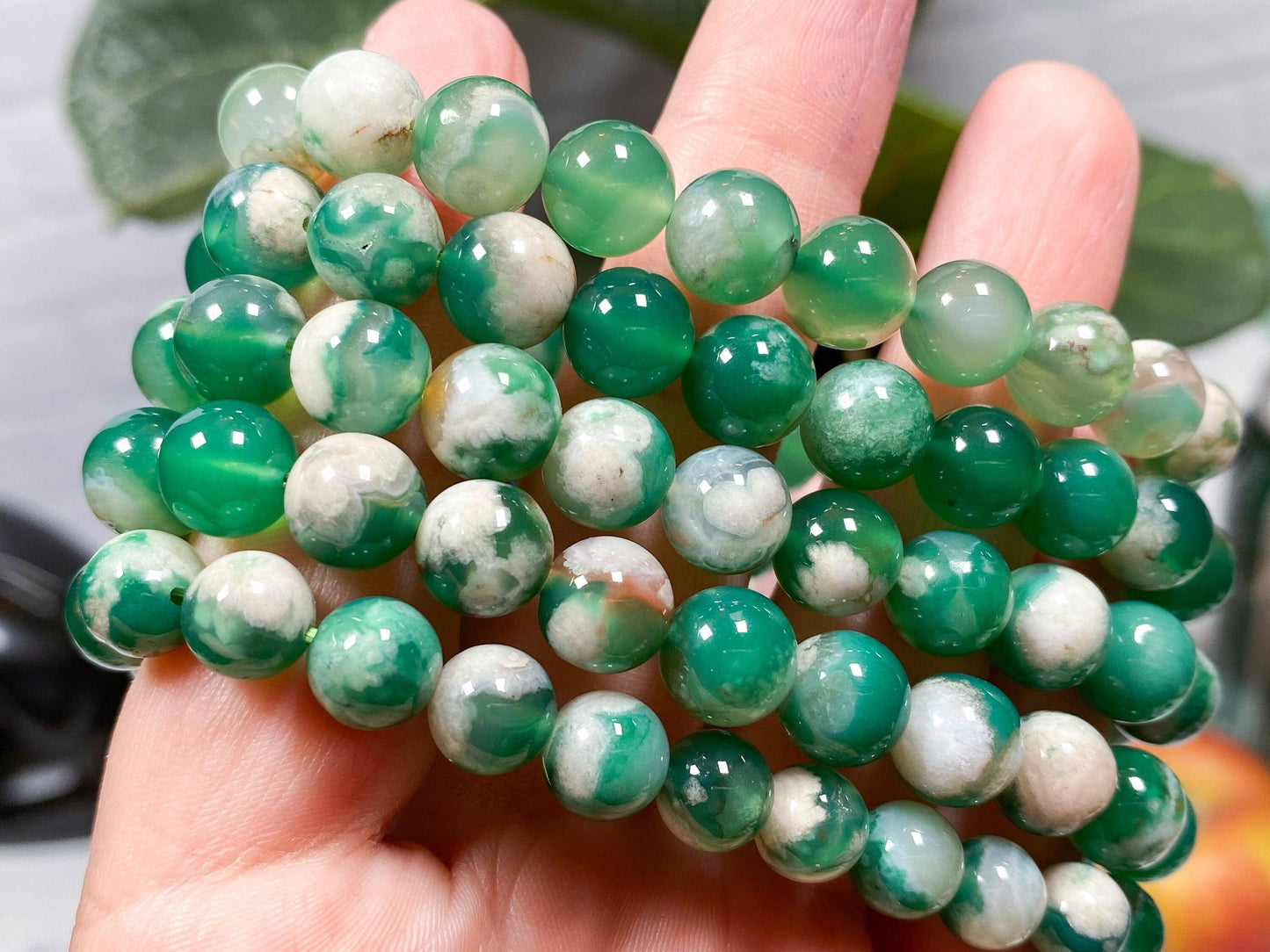 A hand displays the elegant Green Flower Agate Crystal Bracelet by The Crystalary, featuring several strands of polished green and white jade beads. These spherical, smooth, and glossy beads are ideal for heart chakra alignment and exhibit a stunning range of shades from deep green to lighter, almost translucent hues. The background showcases softly blurred greenery.