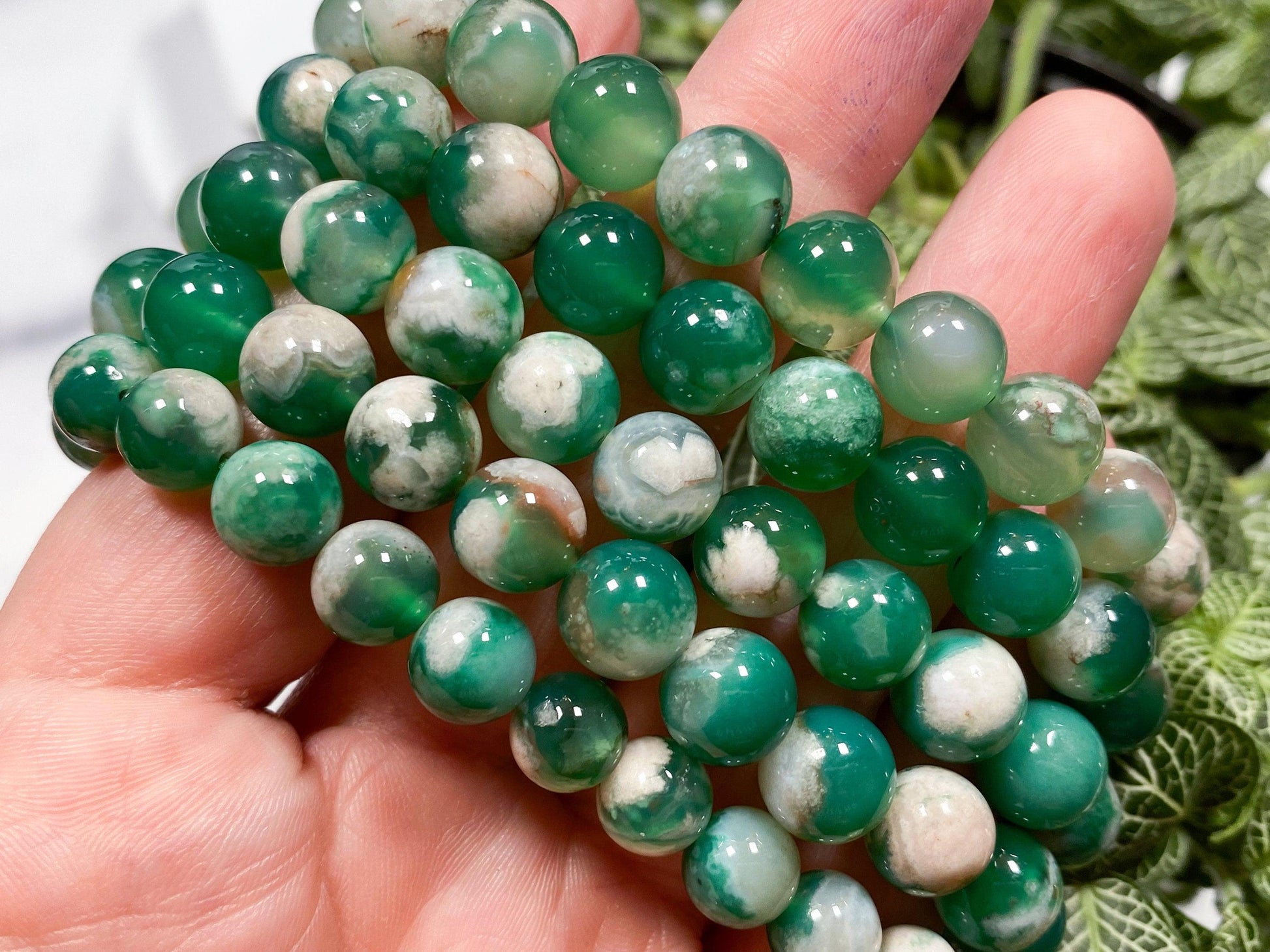 A hand holding strands of round, glossy beads in various shades of green with white marbling. The beads are likely semi-precious stones such as those in "The Crystalary's Green Flower Agate Crystal Bracelet," and are tightly packed together. The background features lush green foliage, evoking a sense of tranquility for the heart chakra.