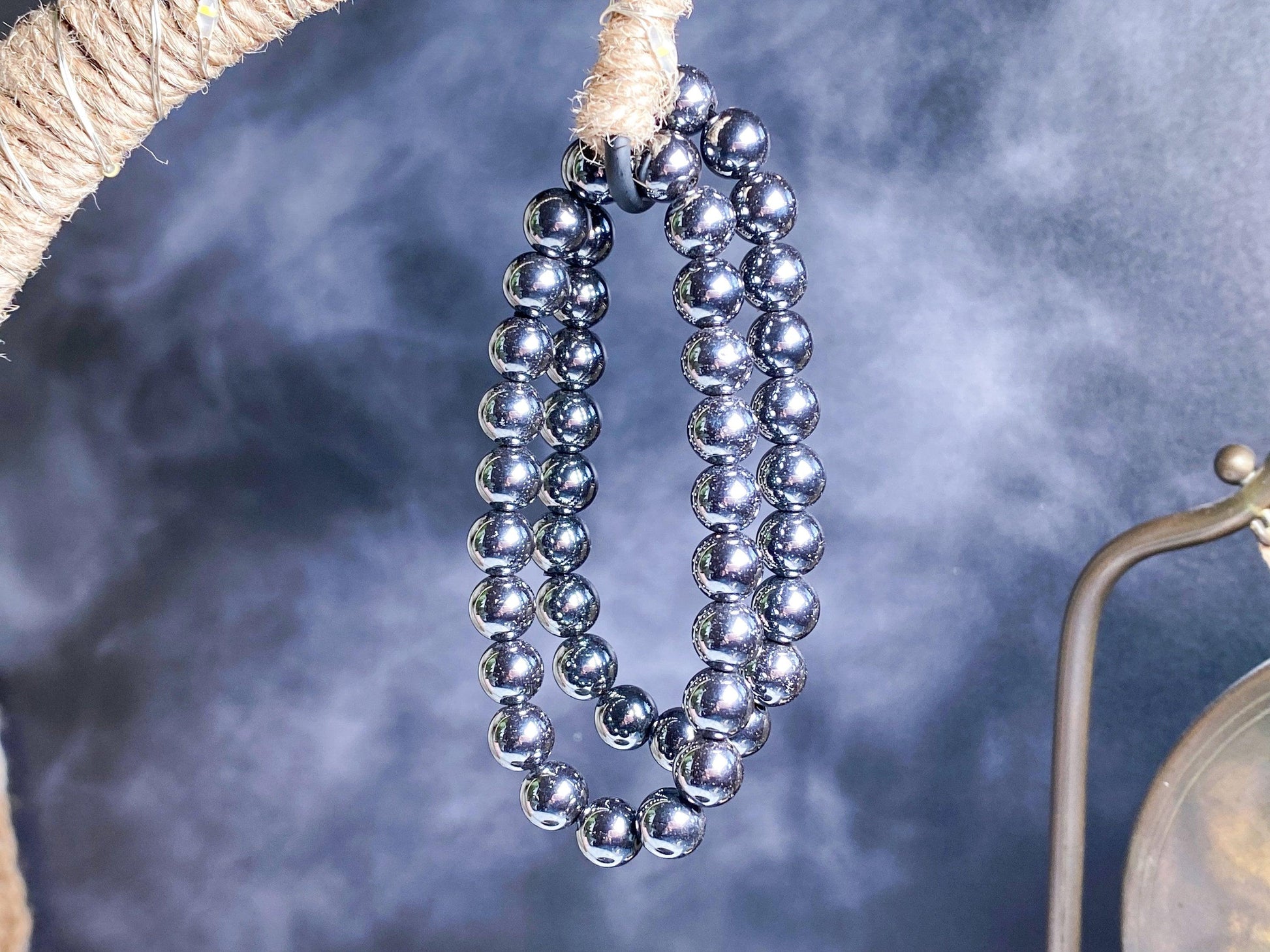 A close-up image of a reflective, metallic, spherical strand showcases the Hematite Crystal Bracelet- 7.5 inch by The Crystalary, hanging from a coarse rope. The smoky or misty background provides a striking contrast to the shiny surface of the bracelets.