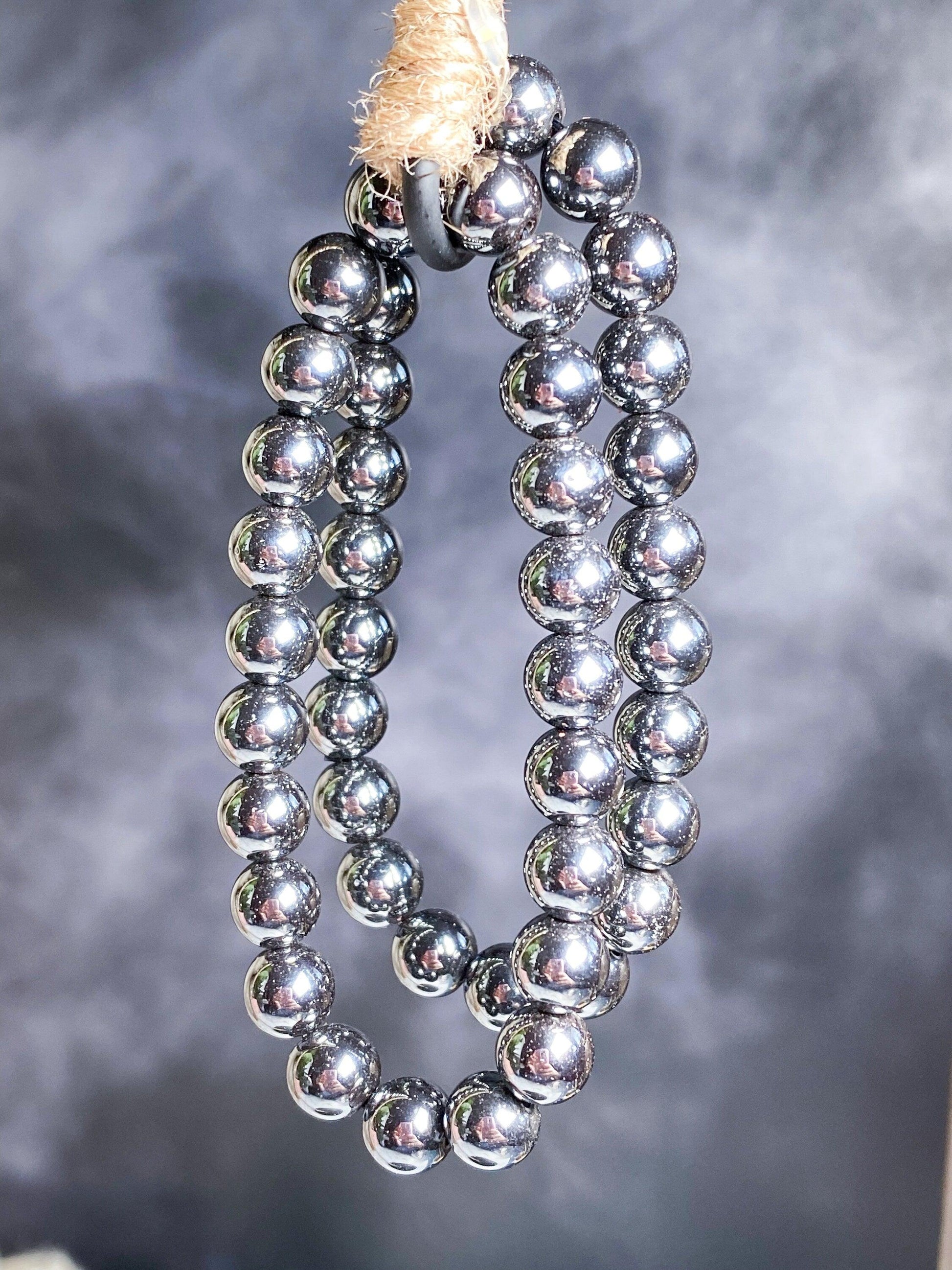 A necklace made of shiny, metallic-looking spherical beads hangs from a textured rope against a blurred, smoky grey background. The reflective surface of the beads gives them a polished and glossy appearance, reminiscent of the stunning Hematite Crystal Bracelet by The Crystalary.