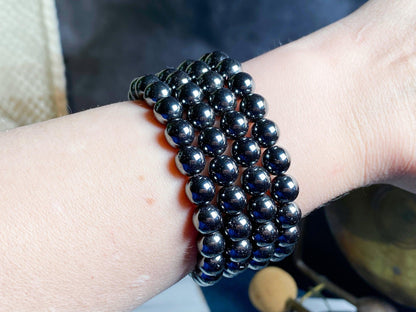 A close-up view of a wrist adorned with multiple strands of the Hematite Crystal Bracelet- 7.5 inch from The Crystalary. The uniformly spherical, highly polished beads reflect light beautifully, giving a glossy appearance. The background is blurred, focusing attention on these protective gemstone bracelets.