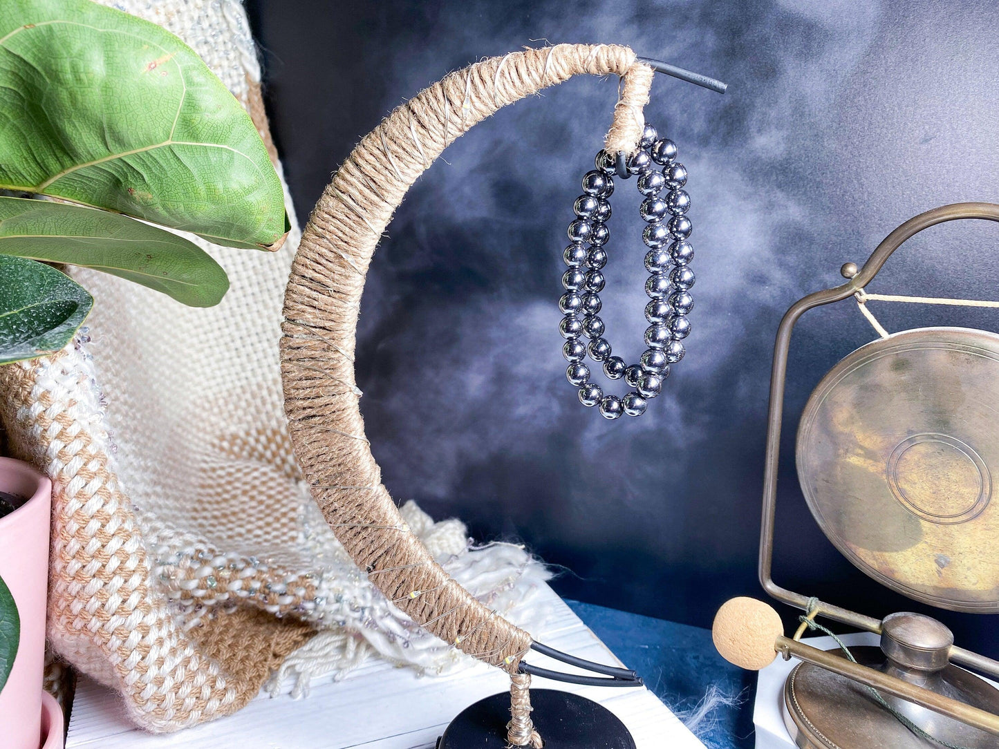 A decorative stand wrapped in twine, shaped like a crescent moon, holds a 7.5-inch Hematite Crystal Bracelet by The Crystalary. Nearby are a gong with a mallet and a large green plant. The background includes a textured beige and white fabric. The scene is misty with a serene ambiance, emphasizing the ethical crystal trade.