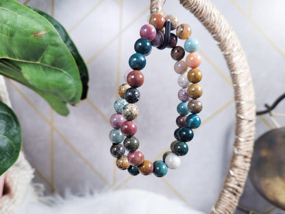 Displayed hanging from a textured hoop is the Ocean Jasper Crystal Bracelet by The Crystalary, a double strand beaded bracelet featuring multicolored round beads, including Ocean Jasper crystals revered for their oceanic healing energy. The backdrop highlights geometric patterns with alternating light and dark shades, complemented by a green plant on the left side.