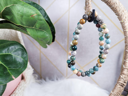 An Ocean Jasper Crystal Bracelet by The Crystalary, featuring a multicolored bead design, is hanging on a rope-wrapped decor piece. The background includes a plant with large green leaves and a wall adorned with geometric tiles. At the bottom of the image, there is fluffy white fabric visible.