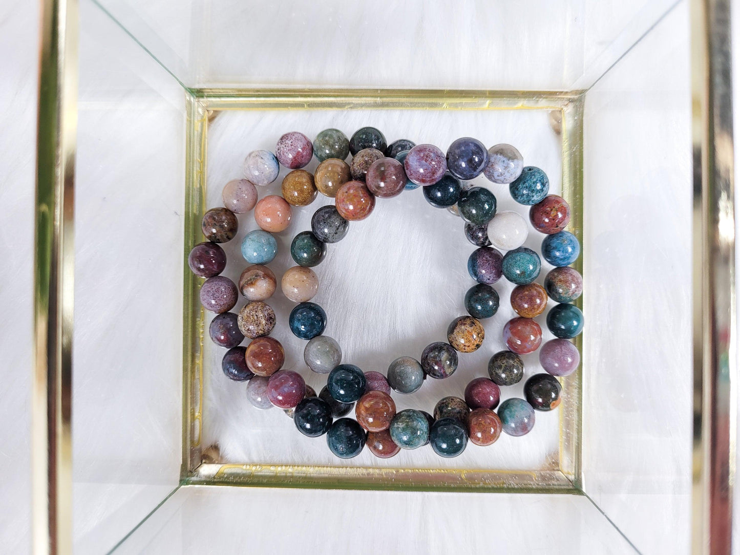 The Ocean Jasper Crystal Bracelet from The Crystalary, featuring colorful, polished gemstone beads in shades of blue, purple, pink, and brown, is elegantly displayed on a light-textured surface within a square glass-framed box. These Ocean Jasper crystals are believed to channel the healing energy of the ocean to open the heart chakra.