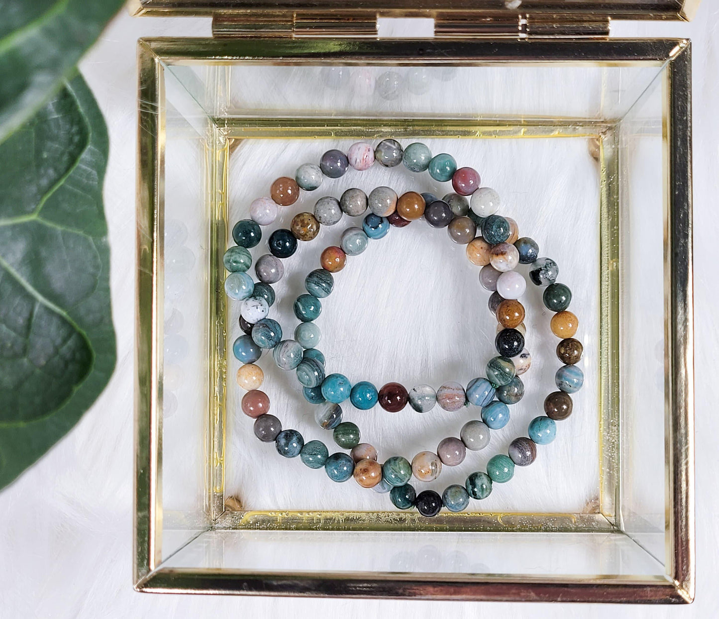 The Crystalary's Ocean Jasper Crystal Bracelets are elegantly displayed in a glass box with a gold frame, resting on a soft, white surface. The multicolored stones of the bracelets showcase an array of hues that resonate with the healing energy of the ocean. A green leaf is partially visible in the upper left corner of the image.