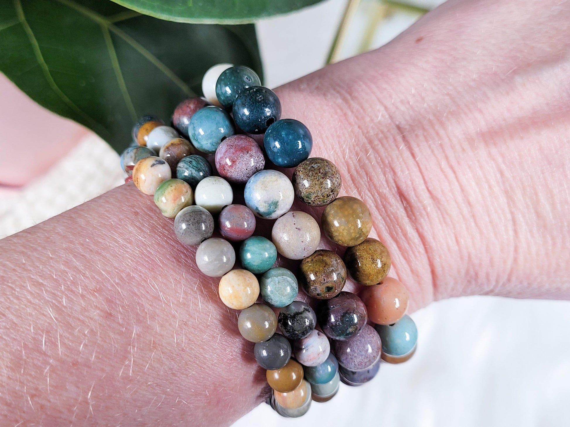 A close-up photo showcases a wrist adorned with several Ocean Jasper Crystal Bracelets by The Crystalary, made of various colorful stones in shades of blue, green, pink, and brown. The stacked bracelets radiate the healing energy of the ocean, with a green leaf visible in the background.