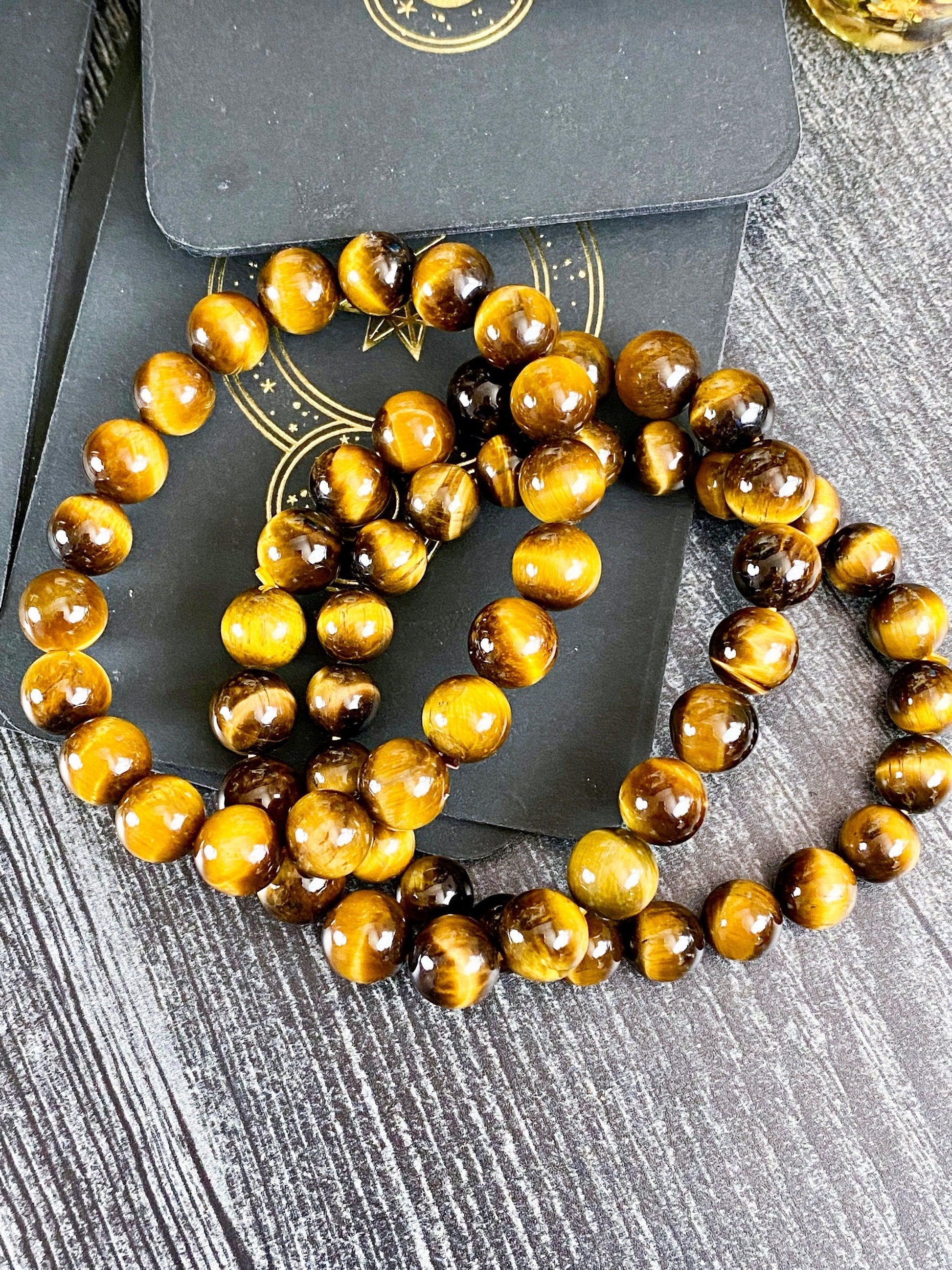 A close-up image of The Crystalary's Tiger Eye Jewelry Crystal Bracelet showcases polished tiger's eye stones. The natural beads exhibit a blend of golden brown and dark brown hues with a reflective, glossy finish. This elegant bracelet is displayed on dark, celestial-themed packaging adorned with star and moon designs.